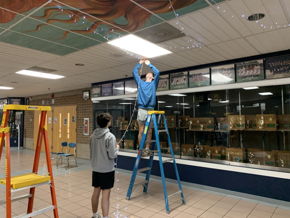 Journalism 1 Seniors Aidan Lawrence and Gavin Mertes set up holiday lights in the hallway leading to the auxiliary gym.